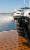 Gloved hand spraying water from the Savwinch Deck Wash Pump nozzle across a boat deck in a marina